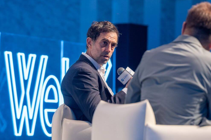 A man in a dark suit speaks into a microphone branded 'CEIBS' while seated at a conference event with blue 'Web3' branding in the background.