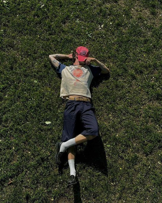 A person wearing a red cap, "Cowboy Shootout" t-shirt, and pinstriped pants lies sprawled on the grass with hands behind their head, embodying the post's "touch grass" mantra—taking a sunny Saturday to literally embrace the outdoors rather than stay glued to crypto charts.