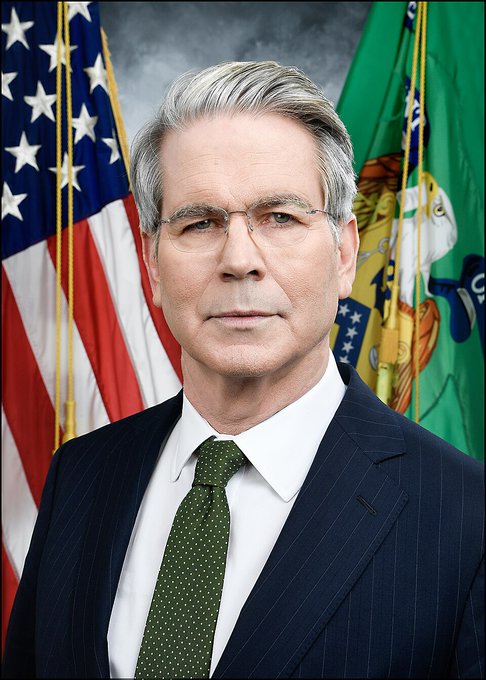 Official portrait of a man with grey hair and glasses, wearing a dark pinstripe suit and green tie, standing in front of an American flag and another official flag.