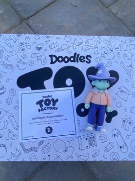 The image shows a "Doodles" x "TOY FACTORY" collaboration box with doodle-patterned artwork and a Certificate of Authenticity, alongside a small wizard figurine wearing a purple pointed hat and green skin. This matches the post, as it appears to be the personal 1/1 Wizard Doodles toy that arrived in the mail.