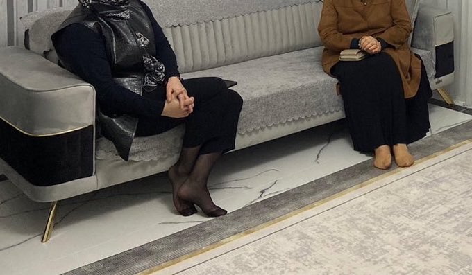Two women sit on a light gray upholstered sofa in a modern interior, one wearing dark pants with patterned tights and a vest, the other in a brown coat over a dark dress, with a marble-patterned floor and area rug visible below.
