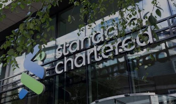 The image shows the exterior signage of Standard Chartered Bank, a financial institution whose logo and name are prominently displayed on a glass building. This relates to the post because Standard Chartered Bank is the entity issuing the bullish Bitcoin price prediction, lending credibility to their call of BTC as "a screaming buy" with a $100,000 target.