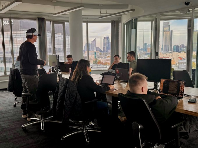 A modern office space with several people working at desks with computers and laptops, featuring floor-to-ceiling windows overlooking a city skyline.