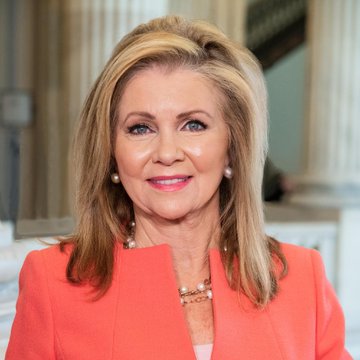 A woman with long blonde hair, wearing a coral blazer and a pearl necklace, smiles in front of a backdrop featuring classical columns. The image accompanies a news update about Senator Marsha Blackburn's comments regarding Democrats voting down the CLARITY Act.