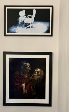 Two framed artworks hang on a light-colored wall: the top shows a white rocking chair in a spotlight against a black background, and the bottom depicts a figure holding a skull.