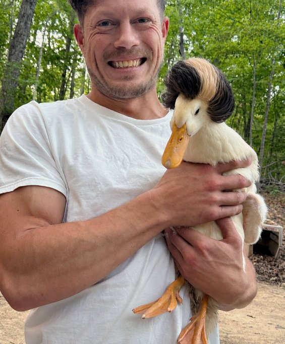 A smiling man in a white t-shirt holds a duck with a distinctive dark patch of feathers on its head while outdoors with green trees in the background.
