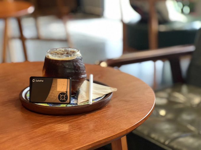A glass of iced coffee sits on a wooden tray on a round table, accompanied by a SafePal hardware wallet, napkins, and a straw, with a blurred indoor setting in the background.