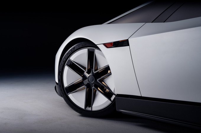 A close-up of a sleek white sports car's front wheel, showcasing a modern multi-spoke alloy rim with black accents and low-profile tires against a dark studio background.