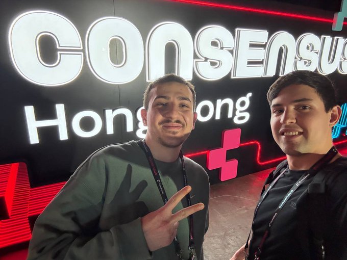 The image features two young men posing for a photo next to a large neon sign that says "Consensus Hong Kong". The man on the left has his hand raised in a peace sign, while the other man stands beside him with his arm around the first man's shoulder. They are both smiling and appear to be enjoying themselves at an event or gathering related to the Consensus community.