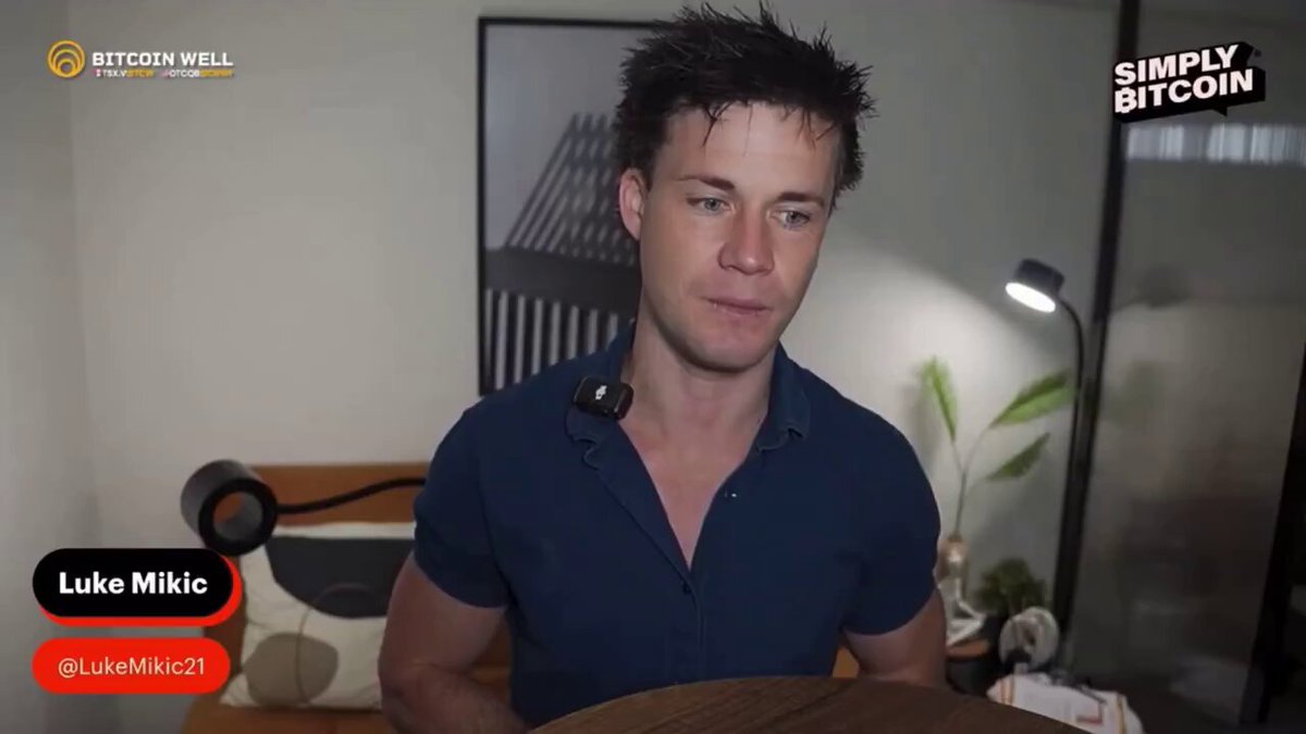 The image shows a man wearing a blue shirt sitting at a desk with a laptop computer, possibly participating in an online discussion about Bitcoin. The background features a picture of a plant and a lamp, adding some visual interest to the scene.