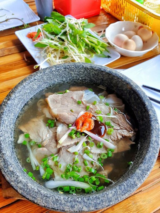 A bowl of pho, a Vietnamese noodle soup, topped with sliced beef, onions, green onions, and chili, served with a side plate of fresh herbs and a bowl of eggs.