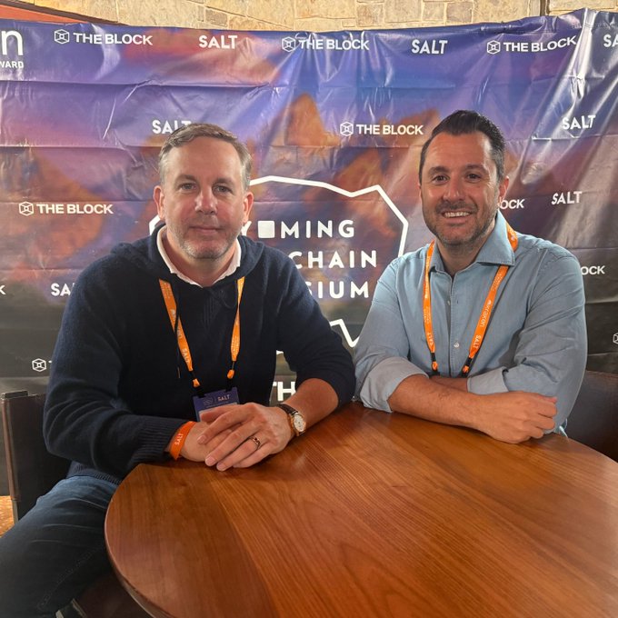 Two men wearing "SALT" lanyards smile while seated at a round wooden table in front of a step-and-repeat banner featuring "THE BLOCK," "SALT," and "COMING CHAIN" (likely "COMING BLOCKCHAIN FORUM") branding, capturing a moment from the on-site interview at the SALT Conference.