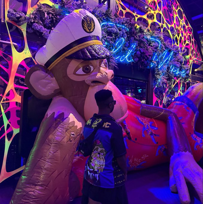 A photograph taken in a neon-lit arcade or themed venue, showing a person wearing a backpack standing in front of a massive monkey mascot statue wearing a captain's hat, with a 'Chill Vibes' neon sign in the background.