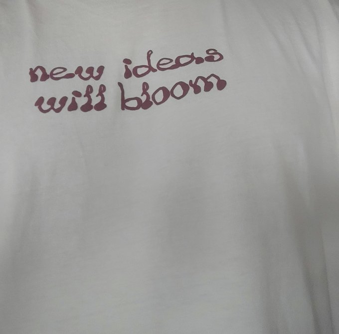A white T-shirt with the dark purple hand-lettered text "new ideas will bloom" printed on the chest. The Arabic post about focusing on cryptocurrencies and required token qualities (real value, institutional adoption, external demand) relates to the shirt's "new ideas will bloom" message, symbolizing optimism that promising crypto investments will flourish.