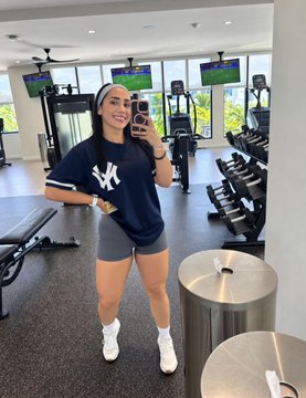 A woman takes a mirror selfie in a gym, wearing a New York Yankees t-shirt, gray shorts, and a white headband, surrounded by dumbbells and treadmills.