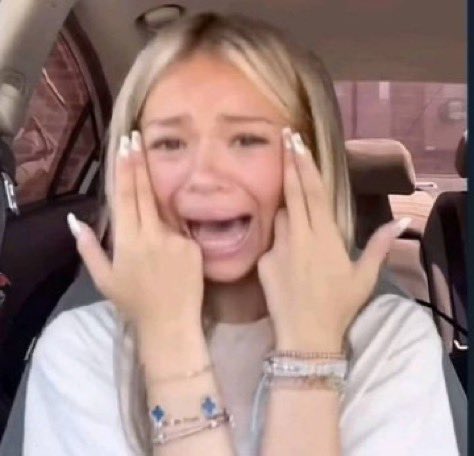 A screenshot of a blonde woman in a car reacting with her hands on her face in an excited, surprised expression, wearing bracelets.