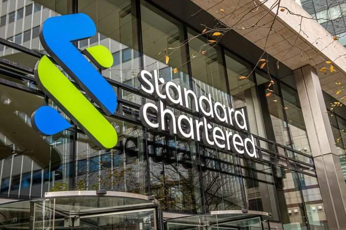 The image shows the exterior signage of a Standard Chartered bank branch, prominently displaying the company's blue and green logo alongside the "Standard Chartered" name in white lettering on the building's glass facade. This visual relates directly to the post, which highlights Standard Chartered's commentary on Strategy's Bitcoin sales and monetization plans.