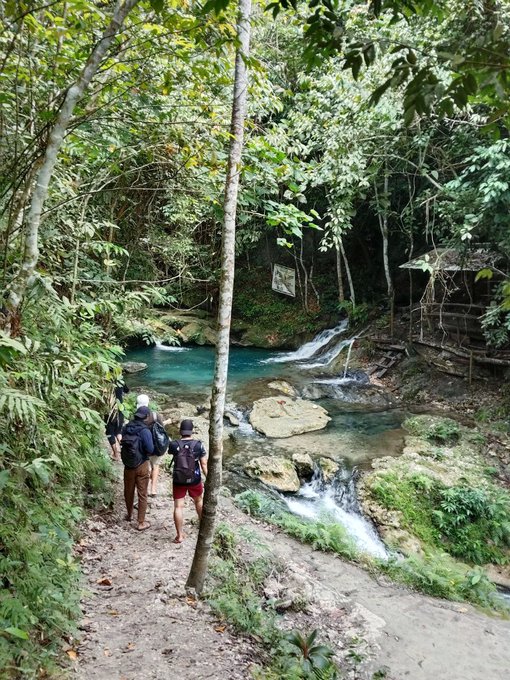 Two hikers with backpacks walk along a dirt trail in a lush green jungle, passing by a small waterfall flowing into a bright turquoise pool.
