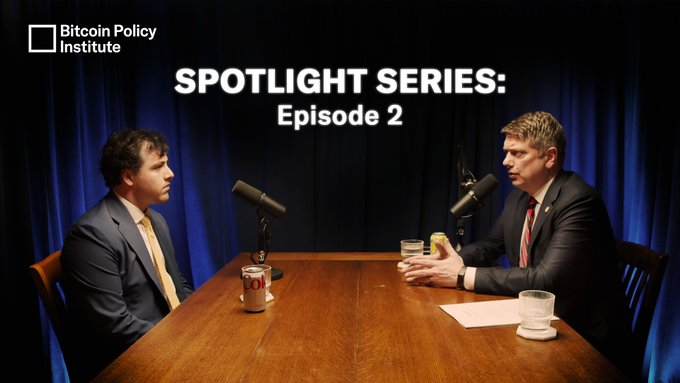 The image features two men sitting at a table with microphones, appearing to be having a conversation or participating in an interview. The background of the image is dark blue, which contrasts with the white text that reads " Spotlight Series Episode 2". There are also some cups on the table, suggesting that they might have been enjoying beverages during their discussion.