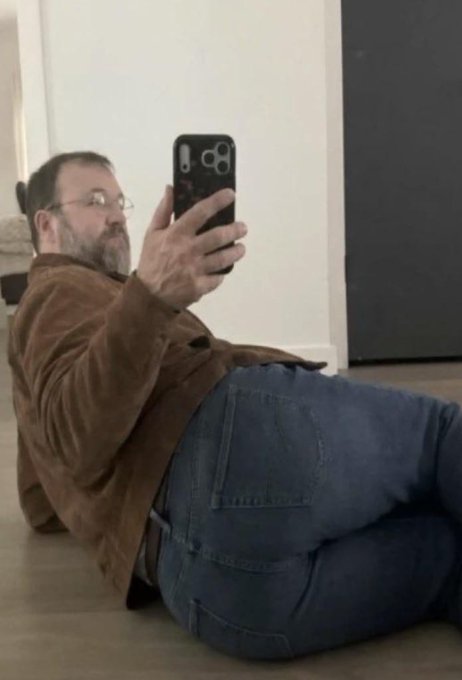 A bearded man in a brown suede jacket and jeans takes a mirror selfie while lying casually on the floor, looking unbothered as he holds up his smartphone. The image plays into the caption by visually conveying the relaxed, confident "aura" referenced—suggesting that despite the ETF news, he's completely at ease and unfazed.