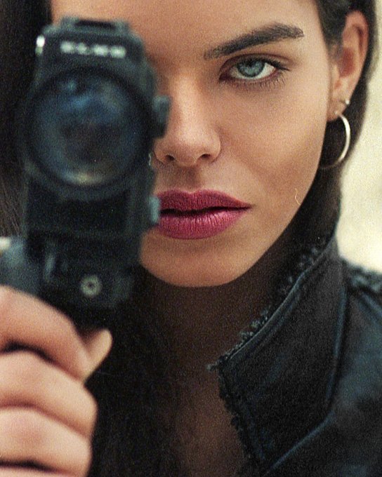 A cinematic close-up of a woman with striking blue eyes holding a gun pointed at the camera.