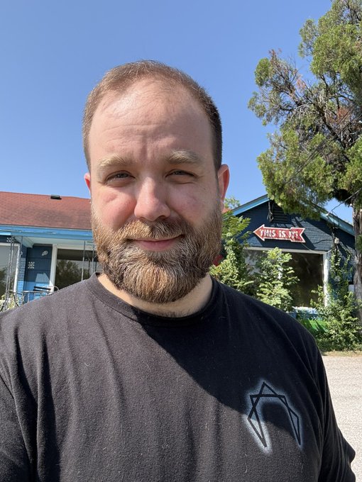 A bearded man wearing a black t-shirt with a geometric logo takes a selfie outdoors, with a blue building, trees, and a 'THIS IS IT!' sign visible in the background.
