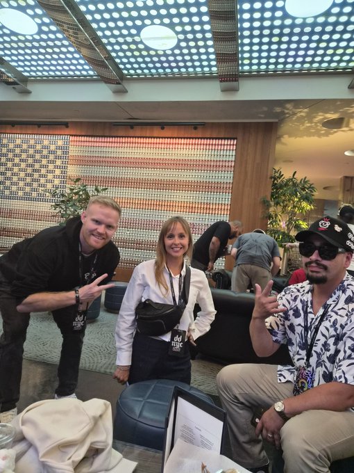 Three people pose in a modern lounge area with colorful beaded curtains; a woman in the center wears a white shirt with a black fanny pack displaying 'dd mic' text.