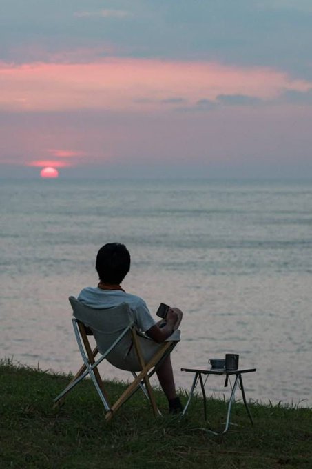 A person sits alone in a camping chair on a grassy cliff at sunset, gazing at the horizon with a mug placed on a small side table beside them.