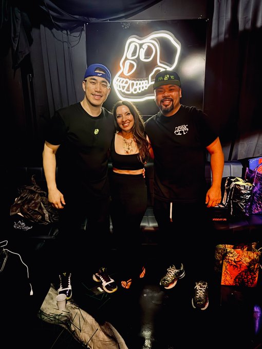 Three people pose for a photo in front of a neon skull sign with 'DOOM' visible, surrounded by a display of sneakers on the floor below them.