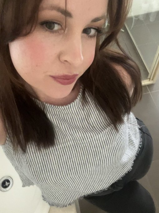 A selfie of a woman with long brown hair wearing a black and white striped sleeveless top, looking directly at the camera.