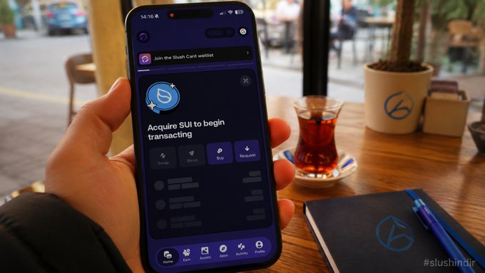 A hand holding a smartphone displaying the Sui wallet app with the prompt 'Acquire SUI to begin transacting', sitting on a wooden café table next to a glass of tea, branded '#slushindir'.