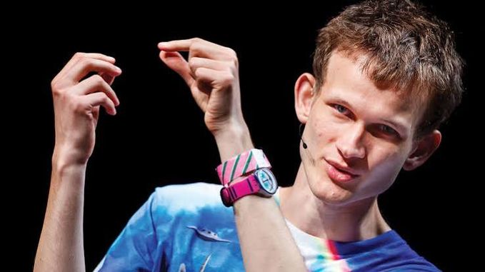 A young male performer with short brown hair and a blue t-shirt is captured mid-gesture on stage, wearing a pink and white striped wristwatch.