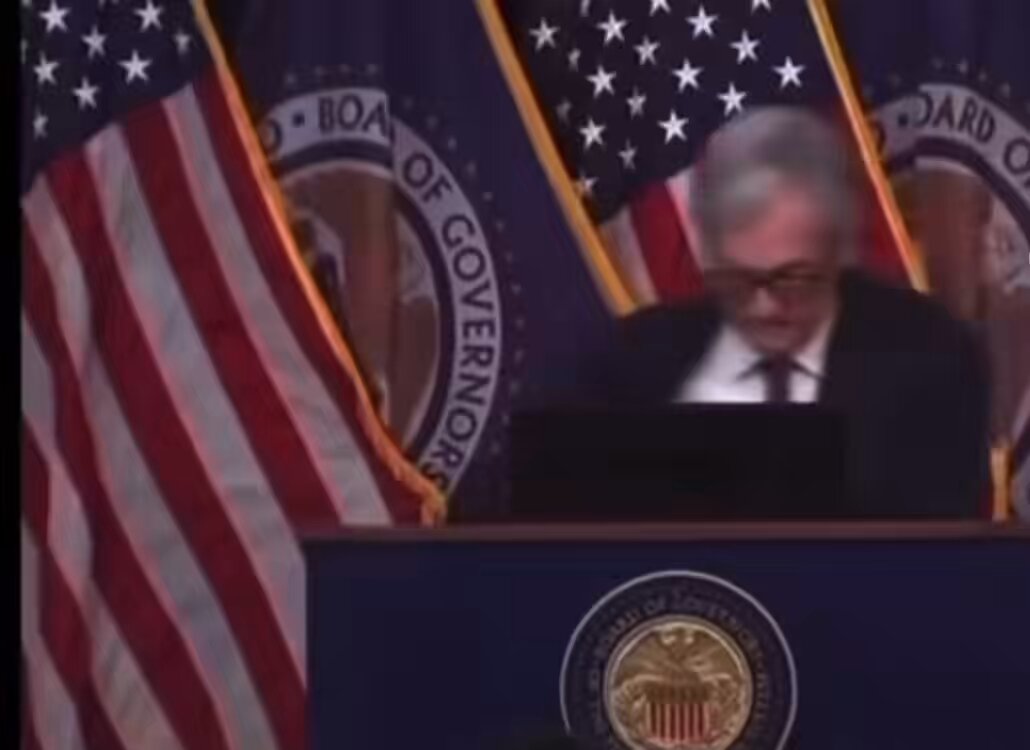 The image features an older man sitting at a podium, possibly delivering a speech or presentation. The backdrop of the scene includes several American flags, with one flag being particularly prominent - the US flag. The text "Today's FOMC will mark the first time in 3,055 days that it won't start with a Jerome Powell" is also visible in the image. This suggests that the event might be related to the Federal Reserve or monetary policy, given the reference to the Federal Open Market Committee (FOMC). The man at the podium appears to be wearing glasses and has a serious expression on his face as he speaks into a microphone.