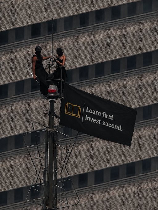Two masked individuals on an elevated platform attached to a building, with one holding a banner that reads 'Learn first. Invest second.' alongside a book icon.