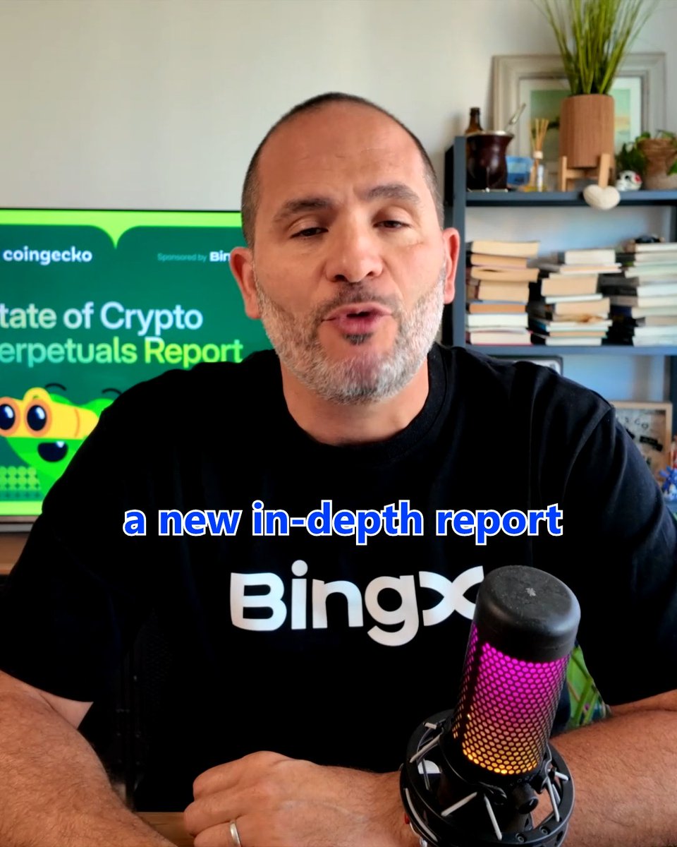 The image features a man wearing a black t-shirt, sitting behind a microphone with a green screen in front of him. He appears to be talking into the microphone or giving a speech. The background includes a TV screen displaying information about the state of cryptocurrency, and there are several books scattered around the room.