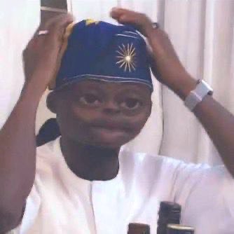 A young boy in white traditional attire adjusts a blue cap with a yellow sun emblem, smiling at the camera.