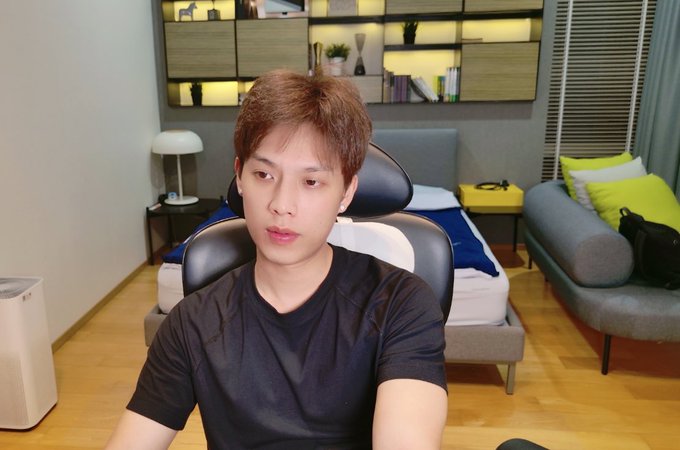 A young man with brown hair wearing a black t-shirt sitting in a gaming chair in a room with a bed, shelving unit, and gray couch with yellow pillows.