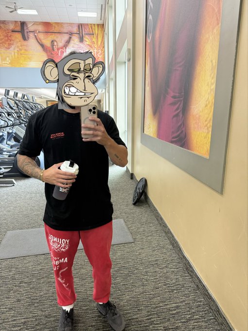 A man wearing a monkey emoji filter takes a mirror selfie in a gym while holding a water bottle and phone.