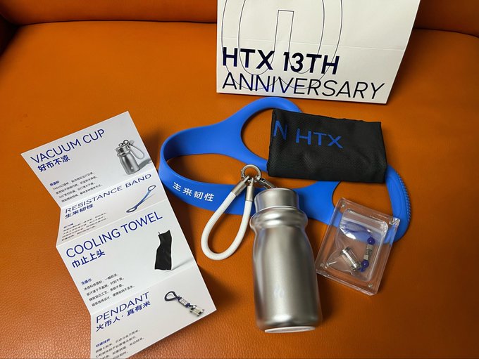 An HTX 13th Anniversary gift box and contents including a vacuum cup, resistance band, cooling towel, and pendant are displayed on an orange leather surface.