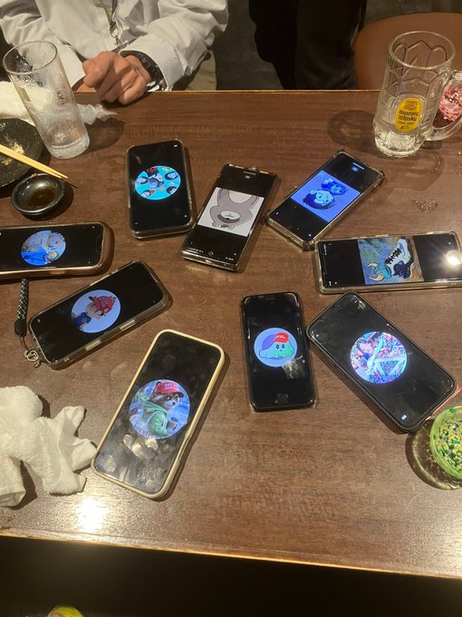 The image shows a wooden table with several cell phones placed on top of it, including one that has a cartoon character on its screen. The table also has various other items such as cups, bowls, and a knife. In addition, there are two people in the scene - one person is sitting at the table while another person is standing behind them. A post with an image of a cartoon character can be seen in the background, suggesting that it might be related to the cell phones on the table or possibly part of a social media post.
