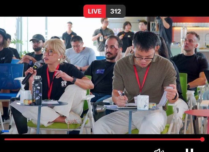 Two judges sit at a table during a live-streamed event, with one woman speaking into a microphone and a man taking notes, while a 'LIVE 312' indicator appears at the top.