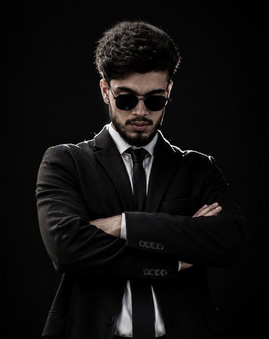 A man with a beard wearing sunglasses, a black suit, white shirt, and black tie, standing with arms crossed against a dark background.