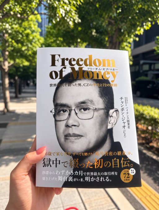 A hand displays the Japanese edition of "Freedom of Money" by Changpeng Zhao (CZ), featuring his black-and-white portrait on the cover, photographed against a sunny tree-lined street outside what appears to be Maruzen bookstore in Japan.