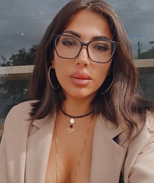 A woman with long brown hair, glasses, hoop earrings, and a beige blazer takes a selfie while wearing layered necklaces.