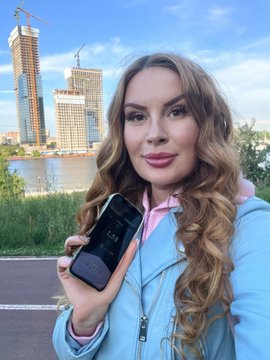 A woman with long wavy hair wearing a light blue jacket and pink top, taking a selfie outdoors while holding up a smartphone, with tall skyscrapers visible in the background.