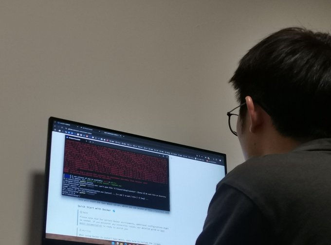 A person wearing glasses and a grey hoodie sits in front of a computer monitor displaying code in a terminal window above documentation for 'Docker'.