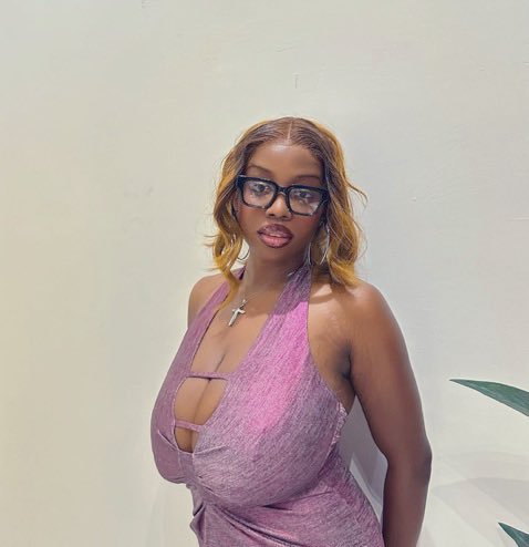 A portrait of a woman with blonde-highlighted hair, wearing black glasses, a cross necklace, and a pink halter dress with a cutout detail.