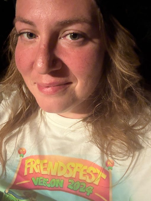 A close-up selfie of a young woman with wavy blonde hair wearing a 'FRIENDSFEST VERONA 2024' graphic t-shirt.