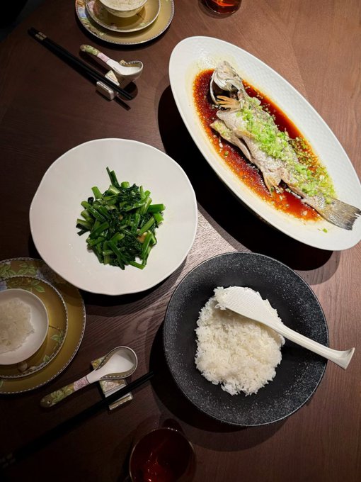 An overhead view of a Chinese meal featuring steamed fish with chili sauce and scallions, stir-fried greens, and white rice, set on a wooden table.