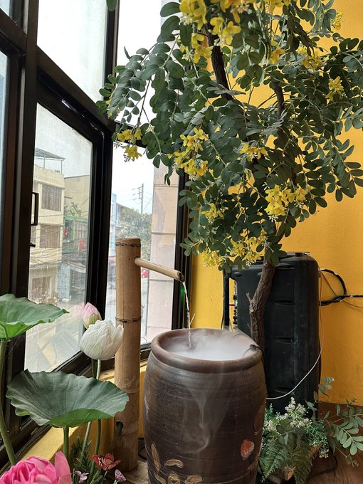 A decorative indoor scene featuring a steaming ceramic pot, a bamboo water feature, yellow flowering branches, pink lotus flowers, and a window showing buildings outside.